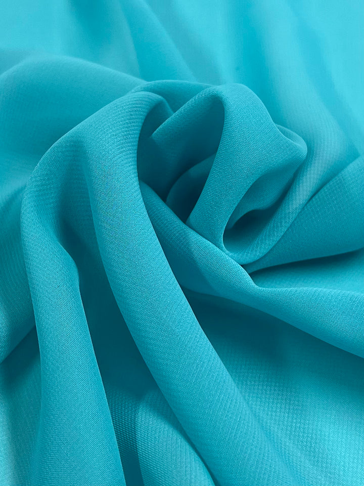 Close-up image of smooth, turquoise Hi-Multi Chiffon - Sea Jet - 150cm by Super Cheap Fabrics with gentle folds and creases, creating a soft and flowing texture. The 100% polyester material appears lightweight and slightly sheer, with subtle variations in light and shadow adding depth to the image.