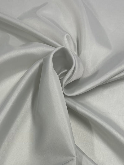 An elegant piece of light grey or silver satin fabric is spread out, creating gentle folds and a centered twist. The smooth, shiny texture of this lightweight fabric reflects light, giving it a luxurious and soft appearance—perfect for lining skirts or as Super Cheap Fabrics' Lining - Celadon Tint - 120cm.
