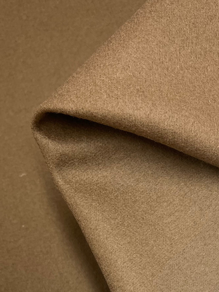 A close-up image showcasing a piece of Melton Wool - Thrush from Super Cheap Fabrics, neatly folded at one corner and lying on a flat, matching surface. The camel brown fabric appears soft and slightly fuzzy, indicative of its 100% wool composition, commonly used for coats and jackets.