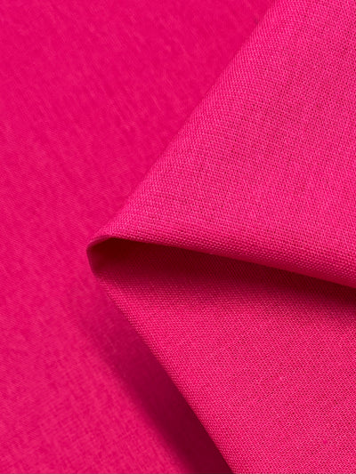 A close-up image of a piece of Super Cheap Fabrics' Linen Blend - Hot Pink - 140cm fabric, with a corner folded upwards. The texture appears smooth and slightly woven, showcasing vibrant color and fine detailing of the medium weight fabric.