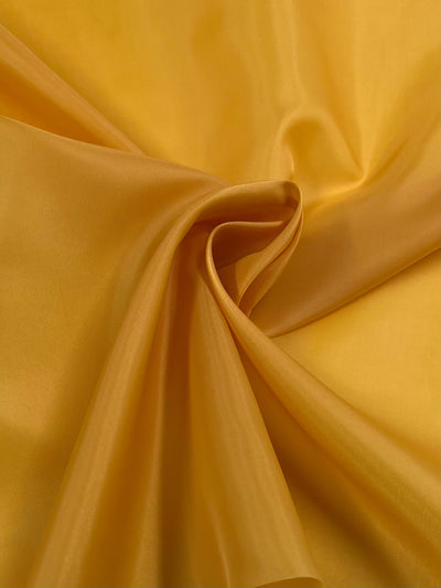 A close-up of vibrant golden yellow fabric, loosely gathered with soft folds and gentle shines, creating subtle shadows and a smooth, silky texture reminiscent of Super Cheap Fabrics Lining - Gold - 150cm.