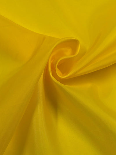 A close-up image of a piece of Super Cheap Fabrics' Lining - Yellow - 150cm, softly gathered in the center to create gentle folds and curves, showcasing its smooth and shiny texture.
